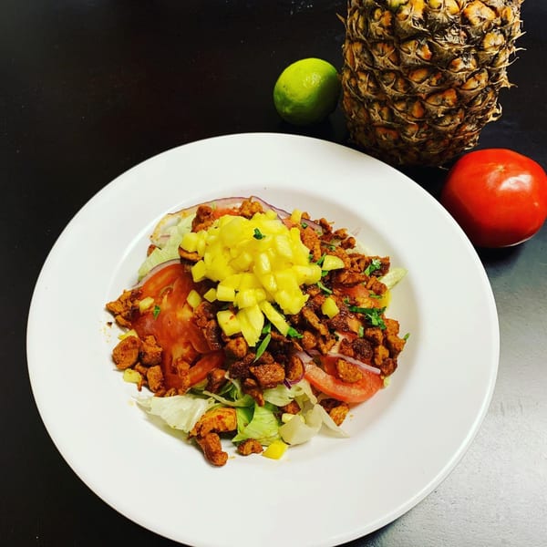 Mexican al pastor salad with pineapple, tomatoes, and lettuce, next to a whole pineapple, lime, and tomato.